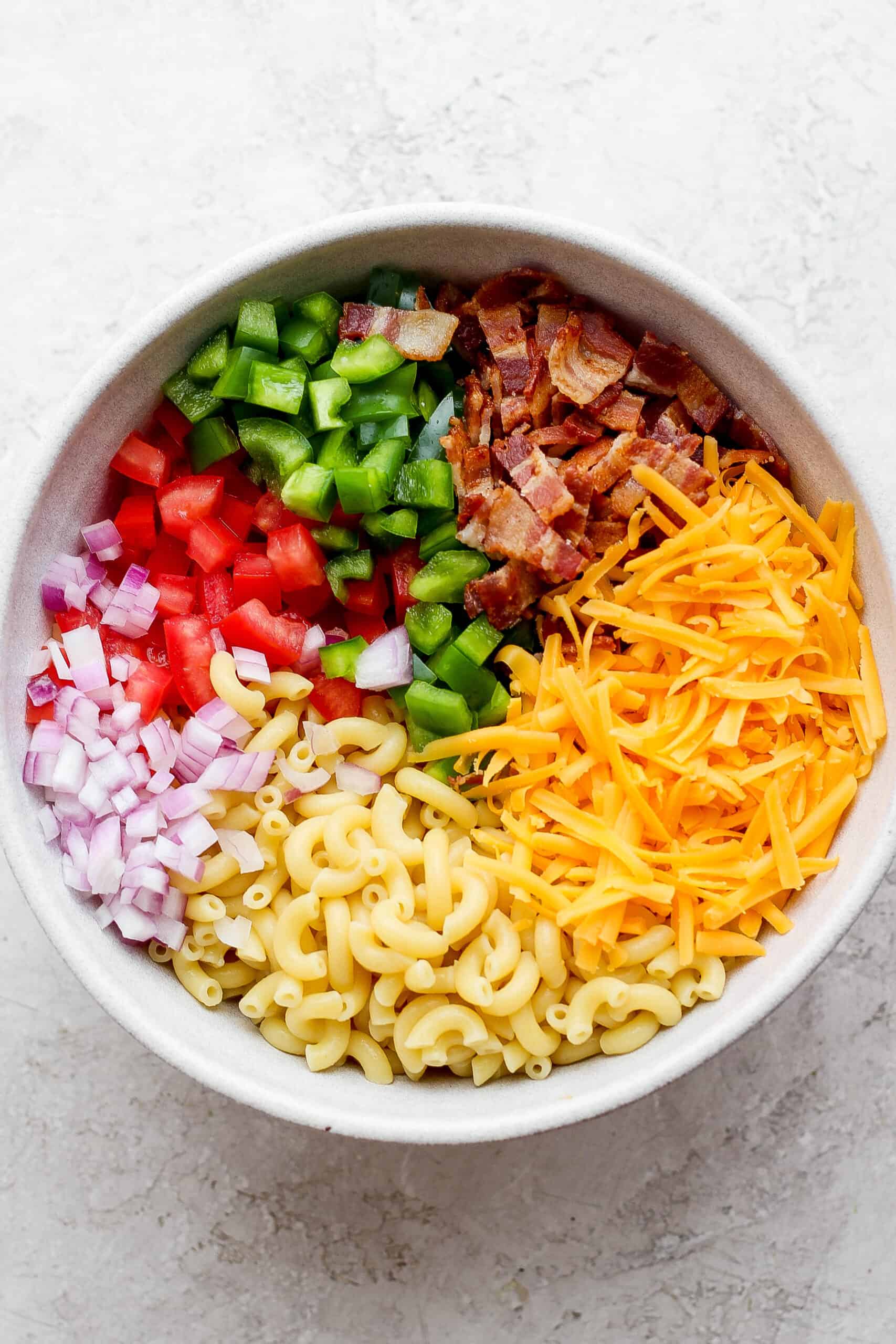 Components of a bacon ranch pasta salad including green onions, red onions, macaroni, cheddar cheese, pepper, and bacon are shown in a white bowl.