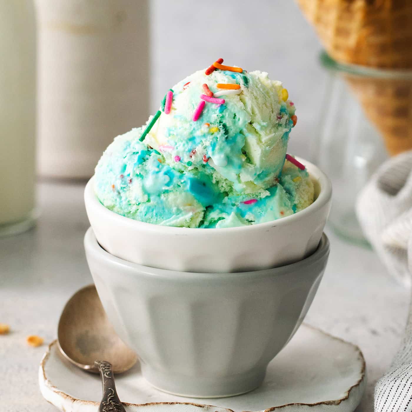 A bowl of blue birthday cake ice cream with a cone of ice cream next to it.