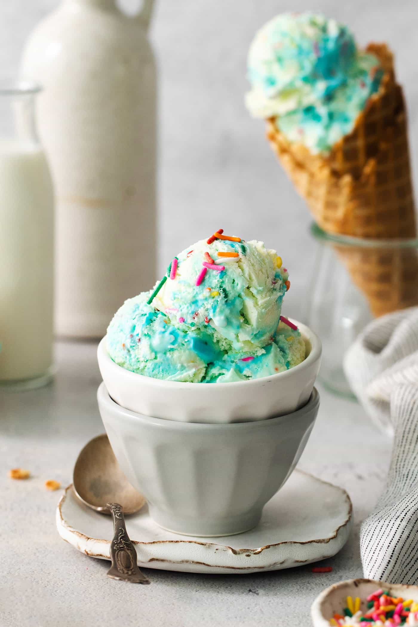 A bowl of blue birthday cake ice cream with a cone of ice cream next to it.