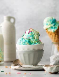 A bowl of blue birthday cake ice cream with a cone of ice cream next to it.