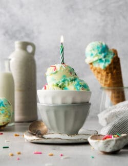 A bowl of birthday cake ice cream topped with a burning candle with a cone of ice cream next to it.