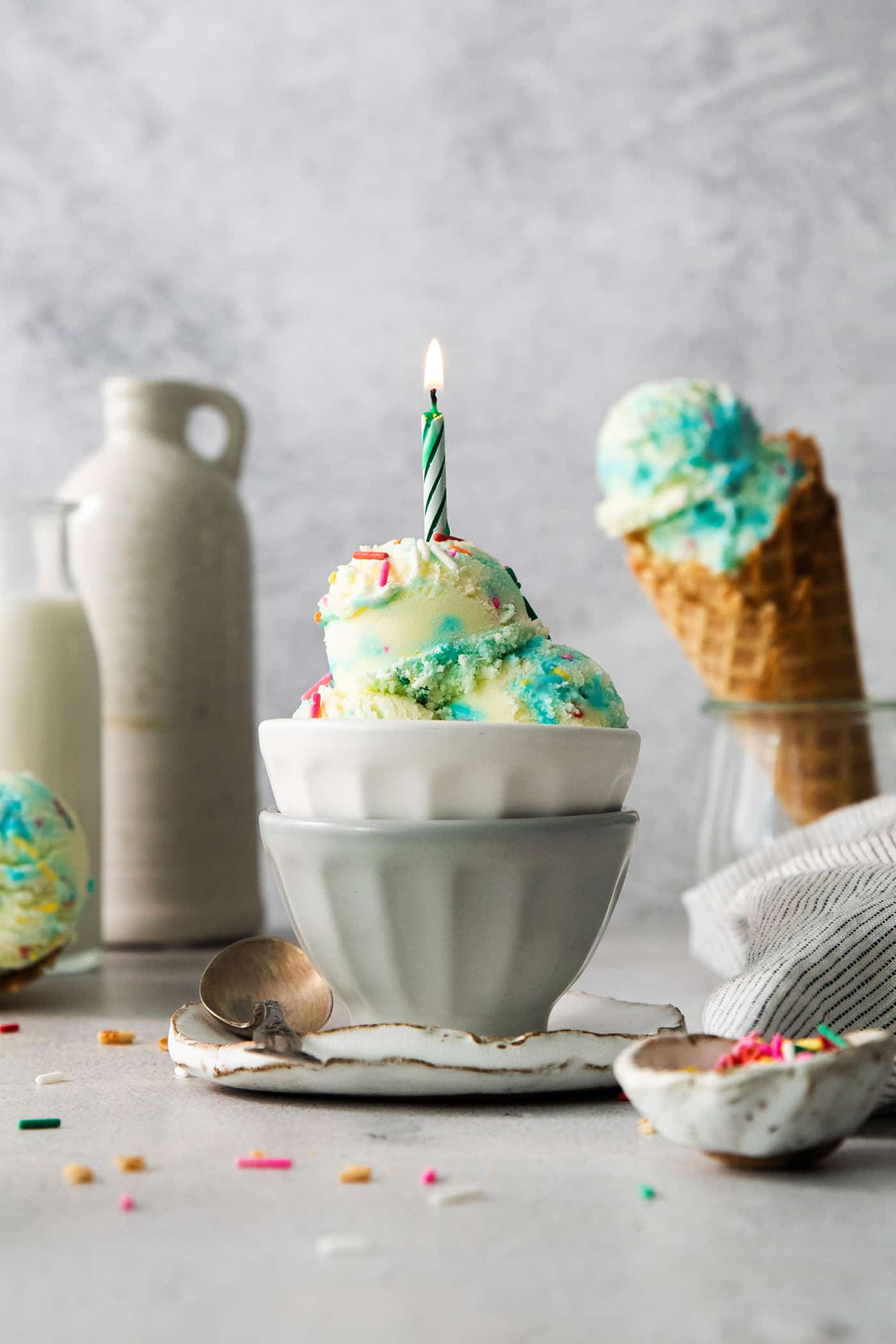 A bowl of birthday cake ice cream topped with a burning candle with a cone of ice cream next to it.