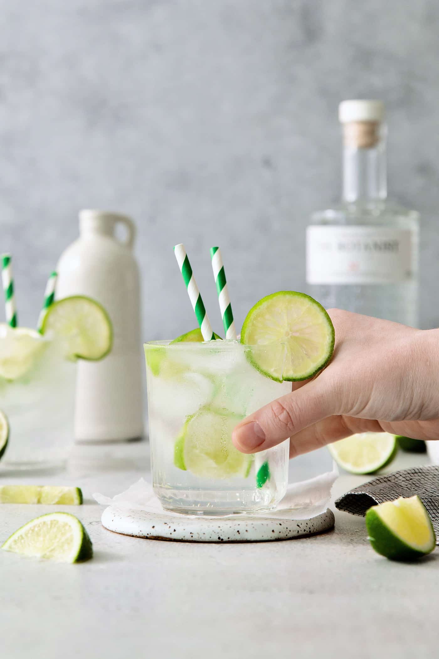 A hand picks up a glass of gin and tonic with two straws and lime circles.
