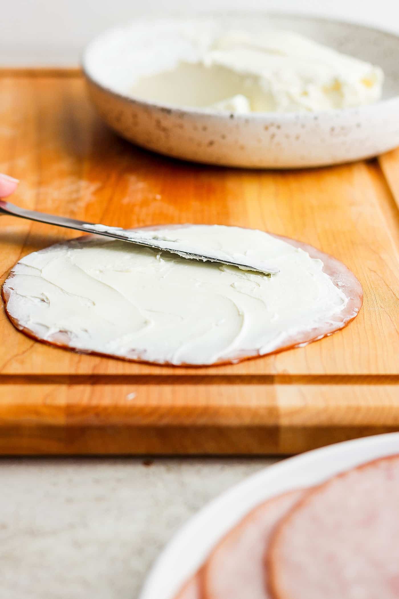 A knife spreads cream cheese on a slice of ham.