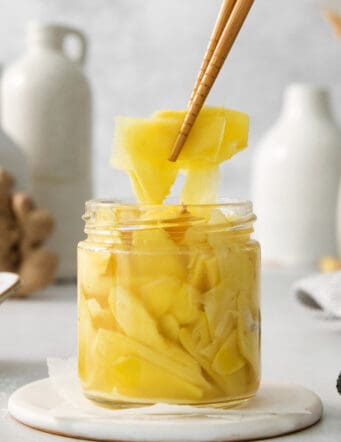 Chopsticks lift out a piece of pickled ginger from a jar.