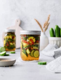 Jars of sweet and spicy pickles with cucumbers in the background.