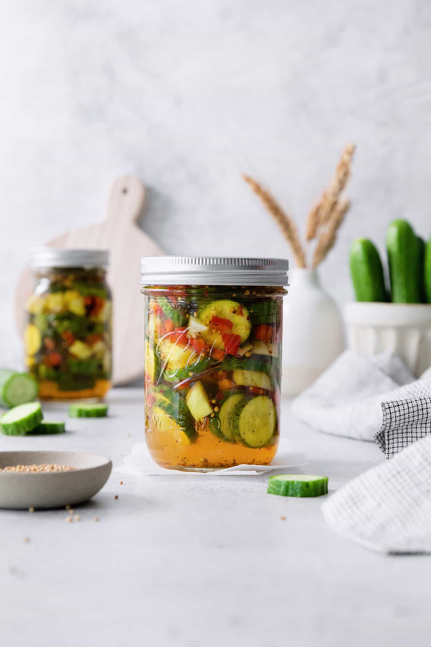 Jars of sweet and spicy pickles with cucumbers in the background.