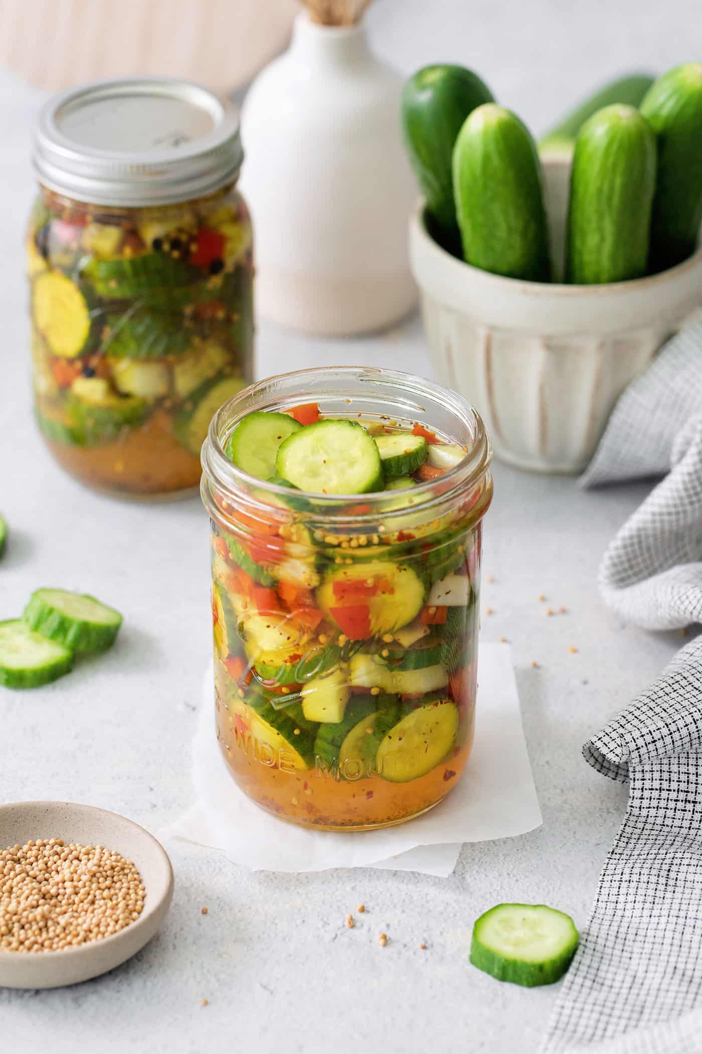 Jars of sweet and spicy pickles with cucumbers in the background.