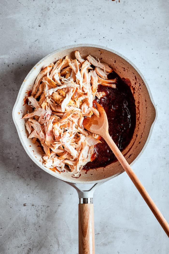 Chicken is stirred into bbq sauce with a wooden spoon.