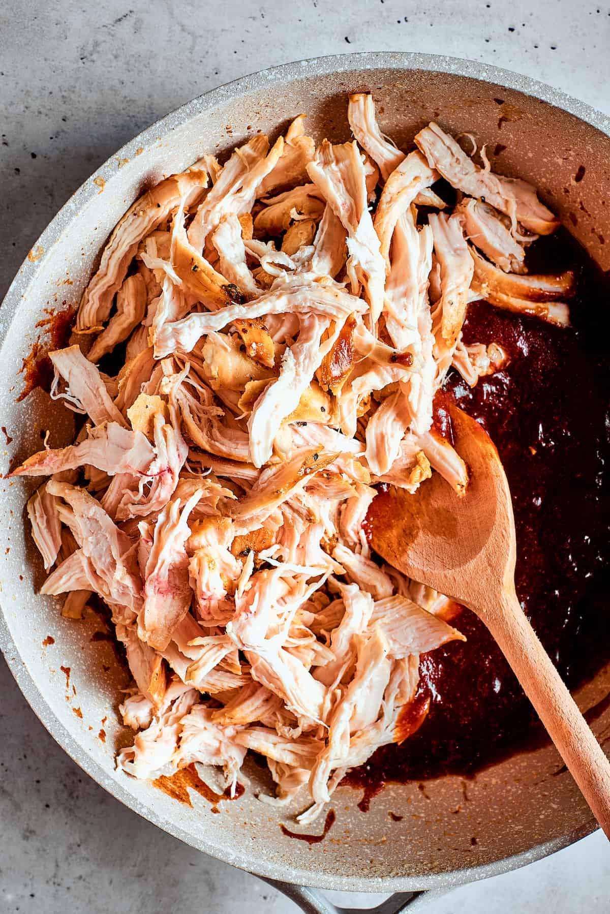 Shredded chicken is stirred into a pot of bbq sauce with a wooden spoon.