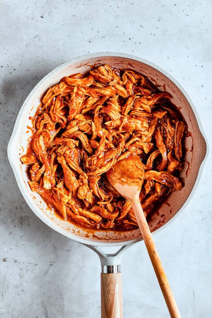 Chicken is stirred into bbq sauce with a wooden spoon.