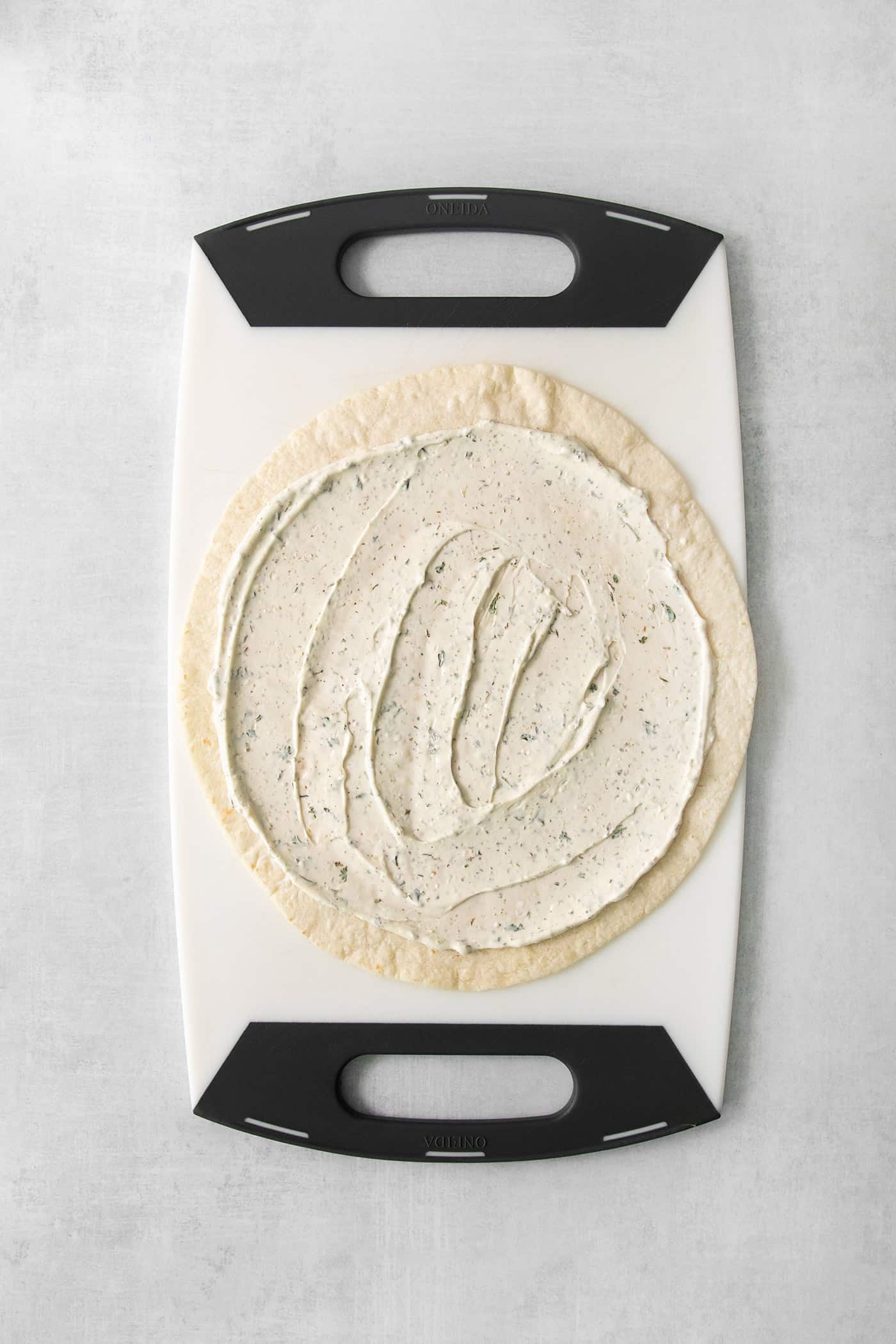 A large flour tortilla on a cutting board is topped with cream cheese spread.