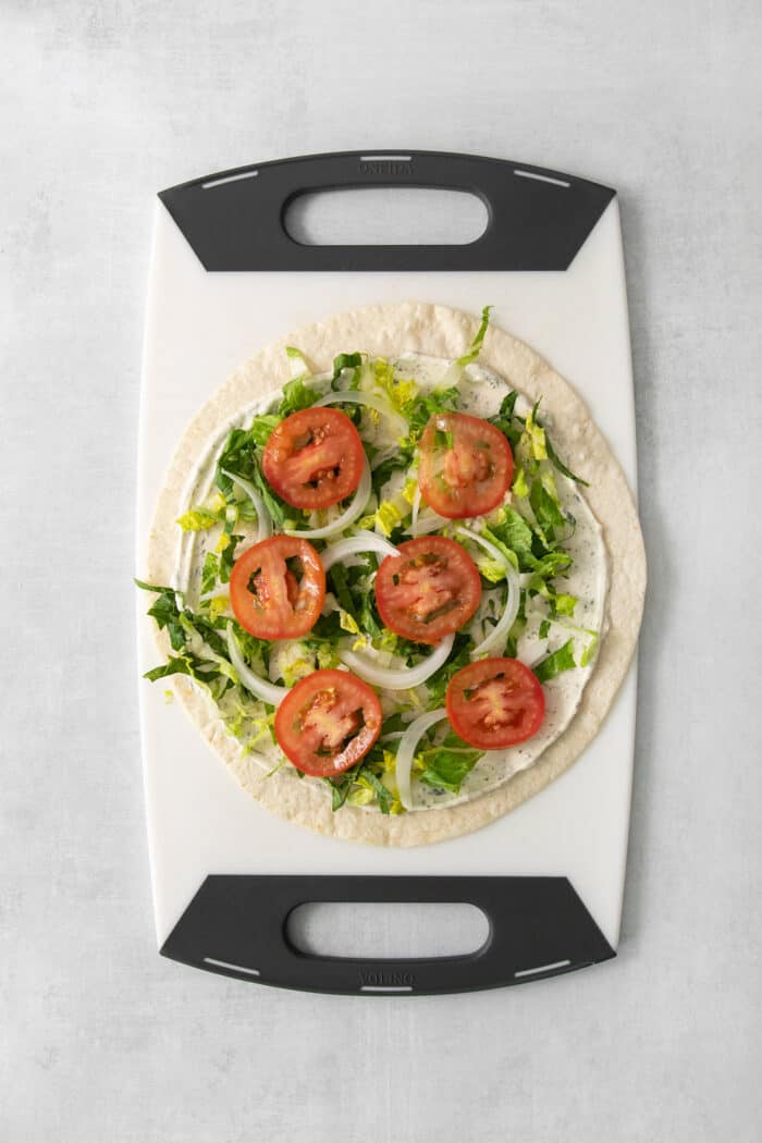 Tomatoes are place atop the lettuce on the flour tortilla on a cutting board.