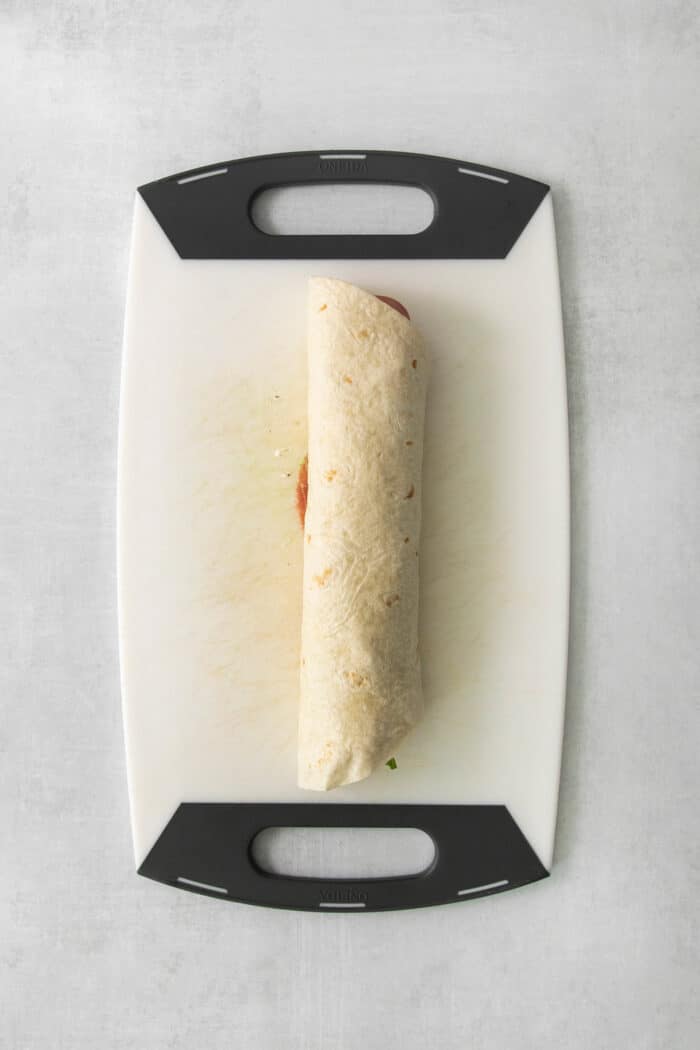 A rolled up Italian pinwheel sandwich is shown on a cutting board.