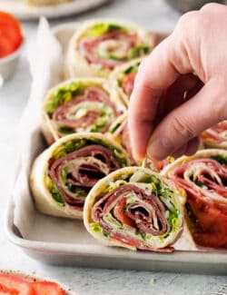 A hand lifts a toothpick holding an Italian pinwheel sandwich.