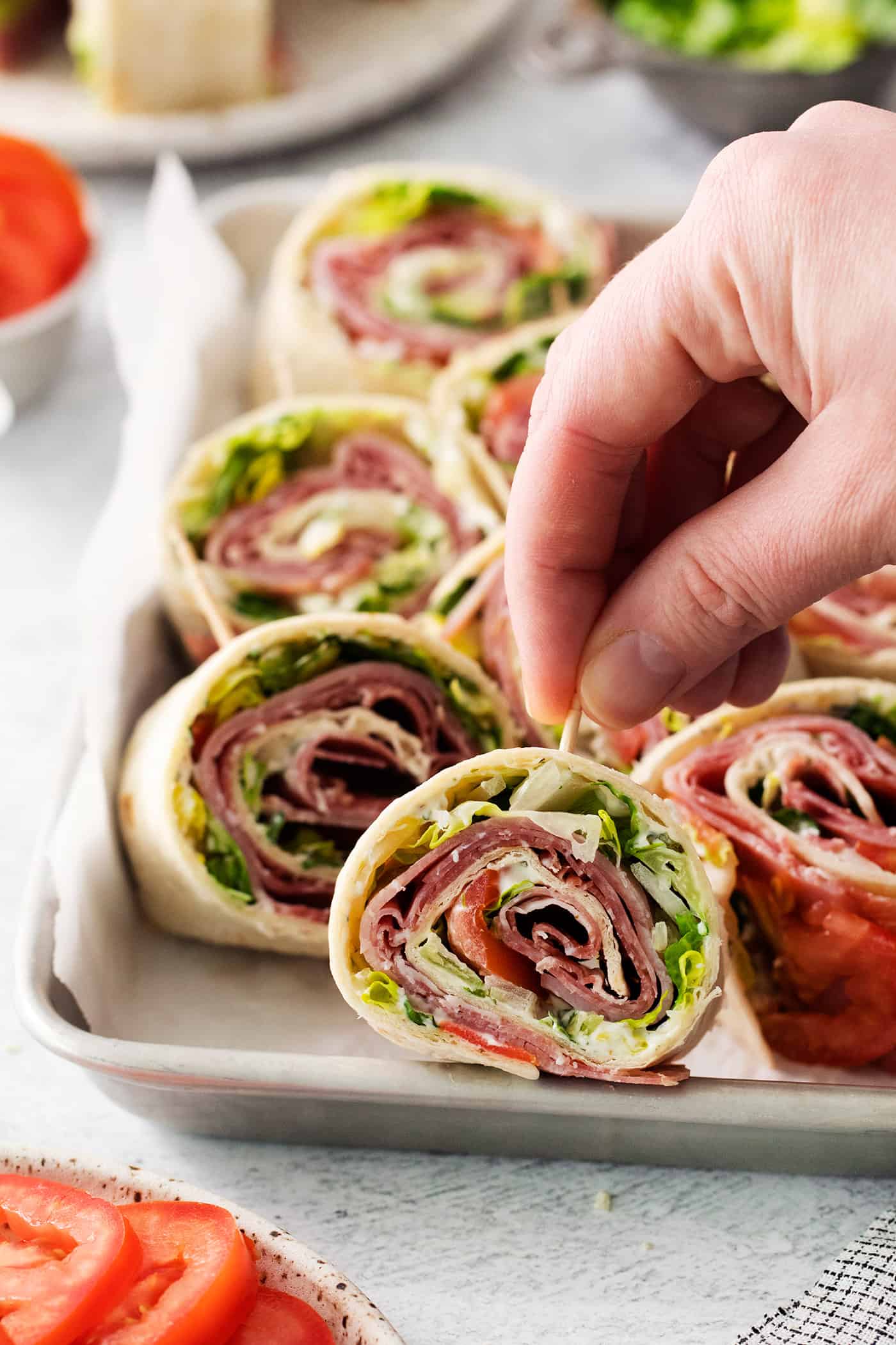 A hand lifts a toothpick holding an Italian pinwheel sandwich.