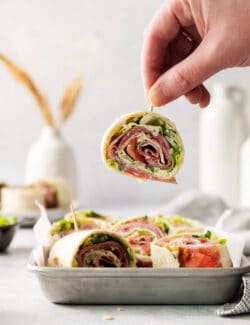 A hand lifts a toothpick holding an Italian pinwheel sandwich.