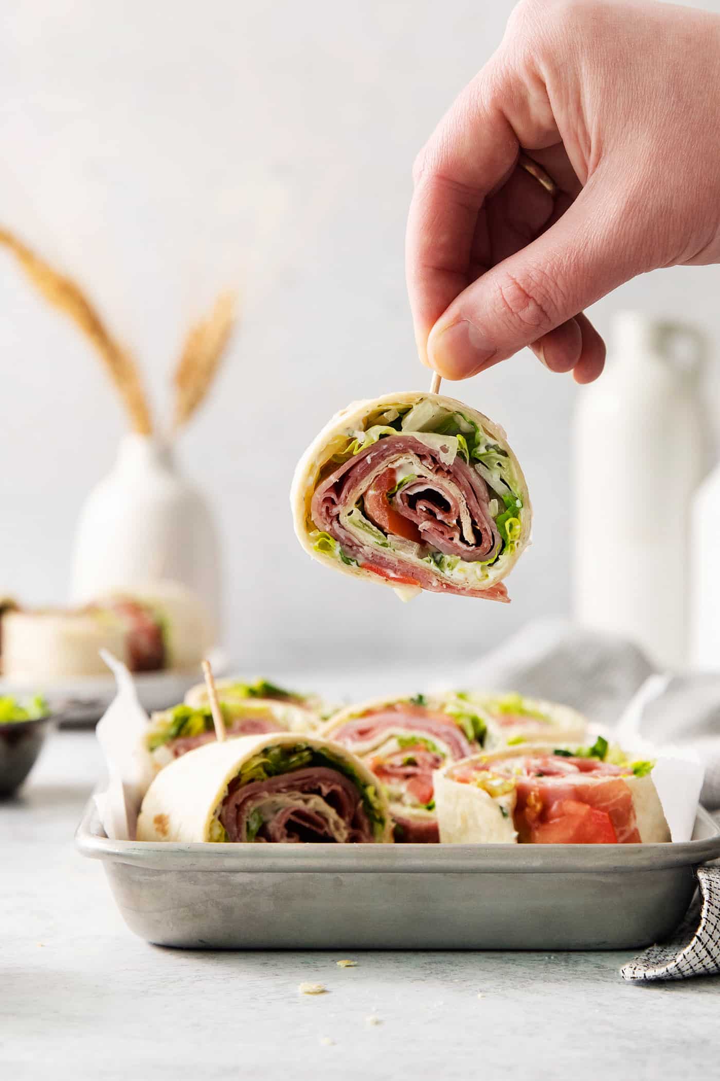 A hand lifts a toothpick holding an Italian pinwheel sandwich.