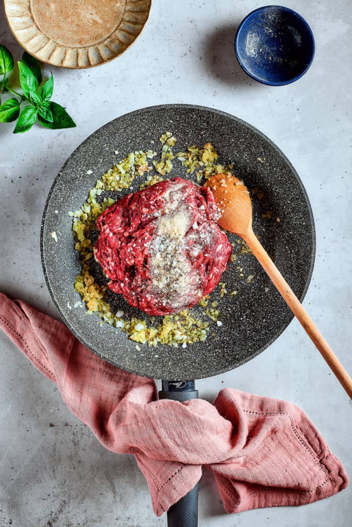Meat is placed in a skillet with spices with a wooden spoon.
