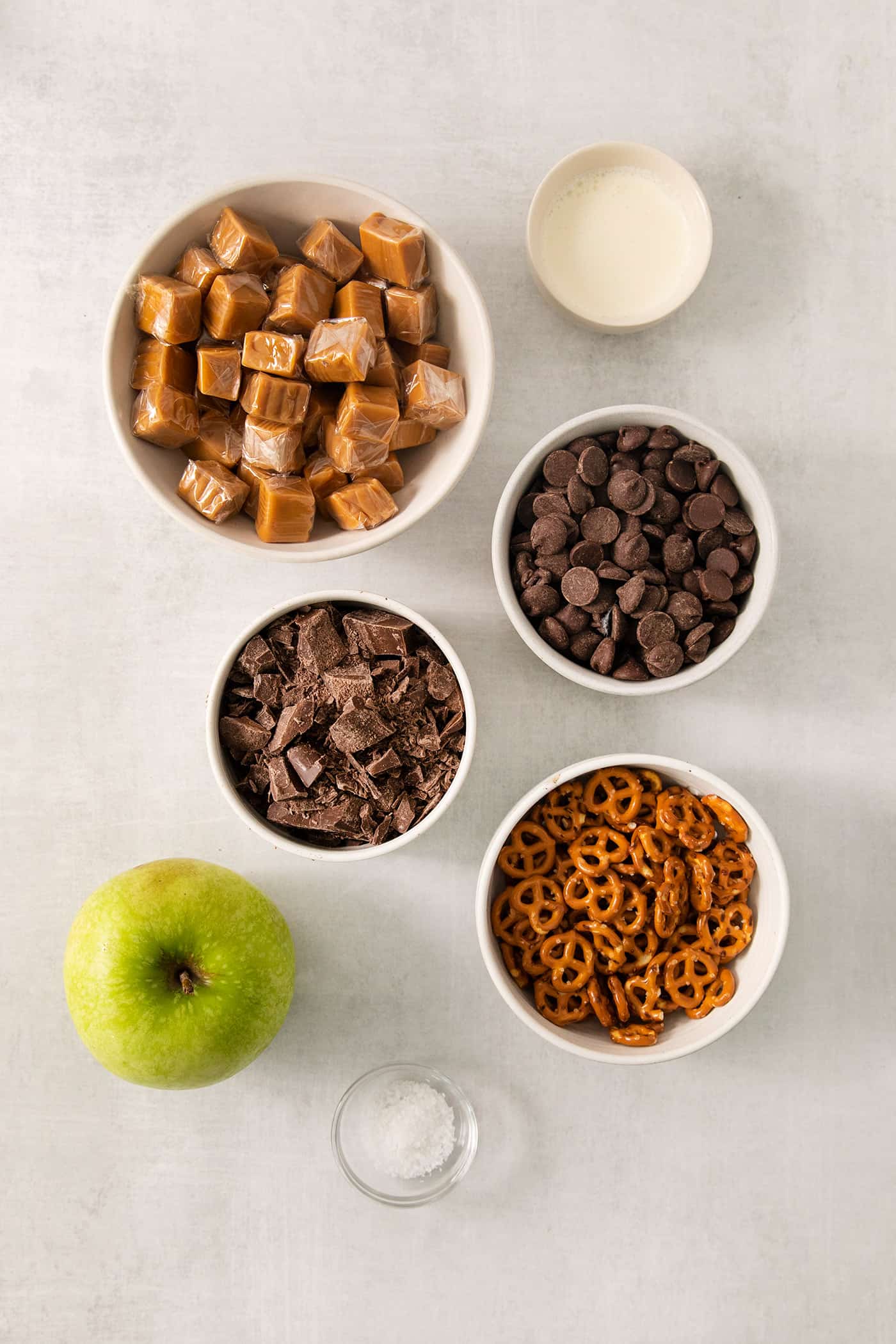 Ingredients needed to make caramel apple bark with pretzels are shown in bowls including caramels, chocolate chips, pretzels, and an apple.