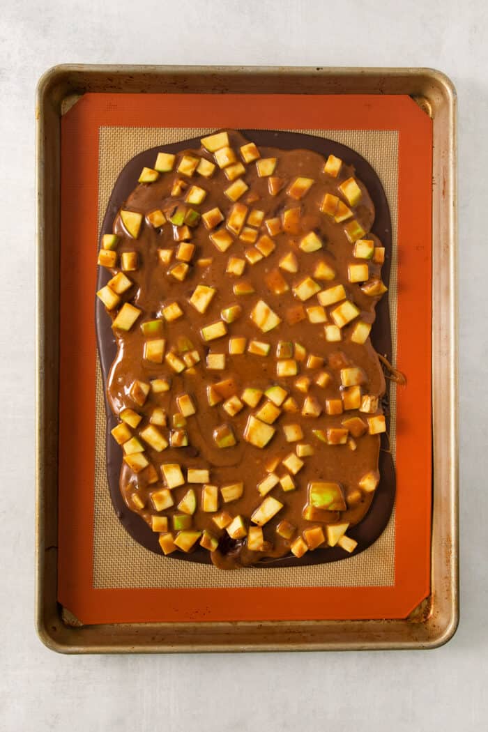 A layer of chocolate is topped with a layer of caramel and apples on a baking sheet.