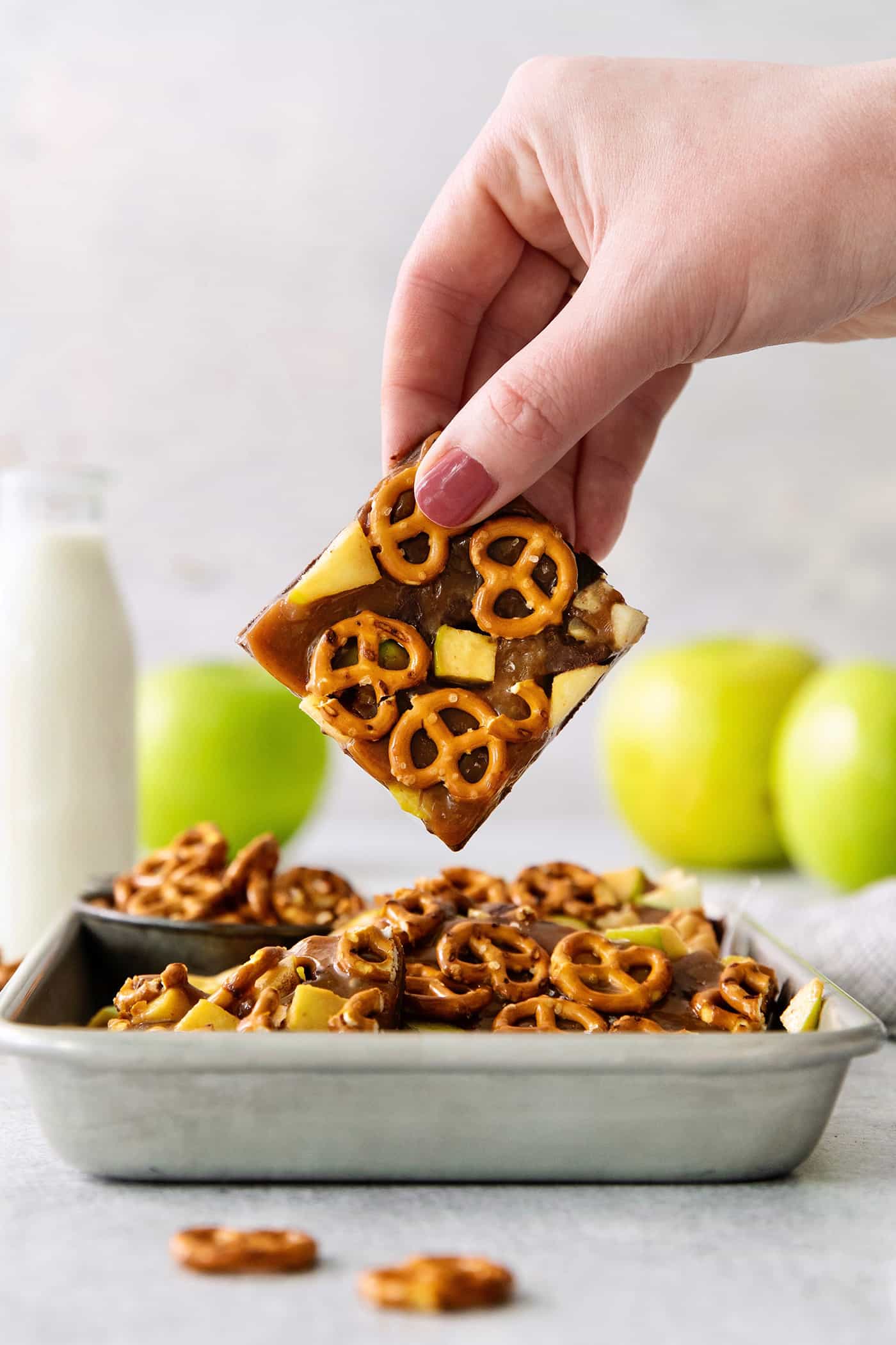 A hand lifts up a piece of caramel apple bark from a try with more pieces of bark with green apples in the background.