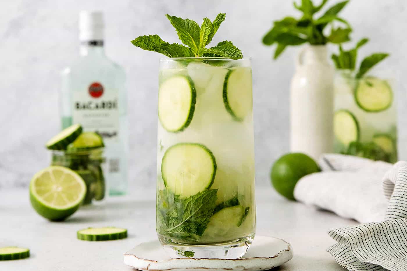 Glasses of cucumber mojito topped with basil are shown with a bottle of rum in the background.