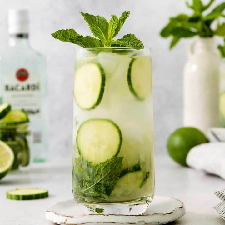 Glasses of cucumber mojito topped with basil are shown with a bottle of rum in the background.