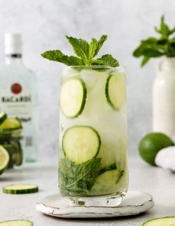 Glasses of cucumber mojito topped with basil are shown with a bottle of rum in the background.