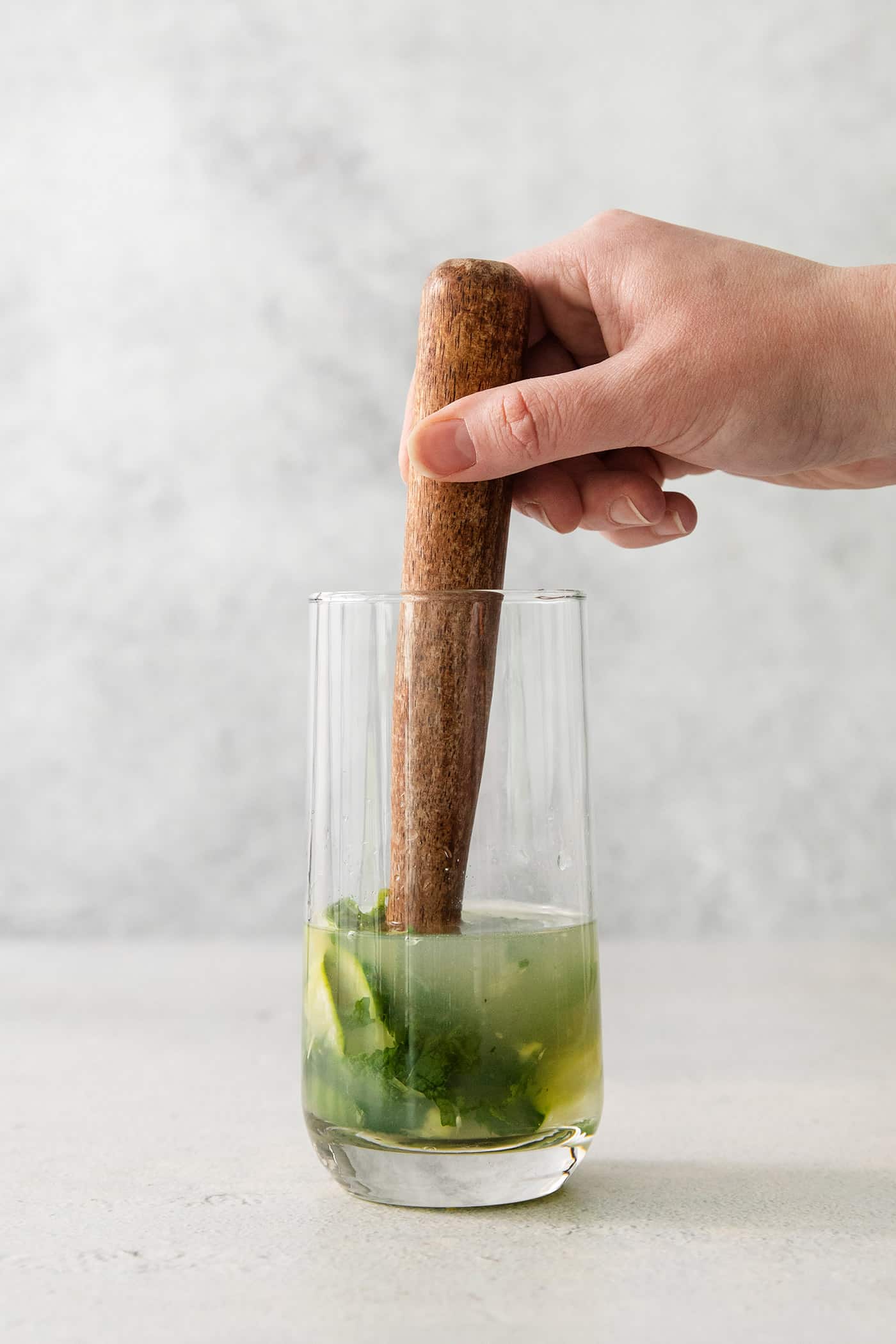 A hand uses a wooden pestle to muddle ingredients for a cucumber mojito.