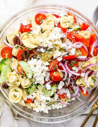 A bowl of Greek tortellini salad is shown on a white table with serving spoons.