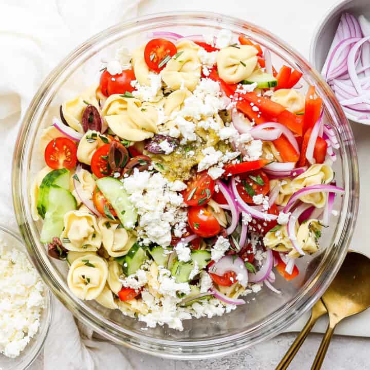 A bowl of Greek tortellini salad is shown on a white table with serving spoons.