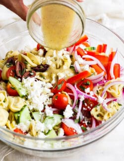 A hand pours dressing onto a bowl of Greek tortellini salad.