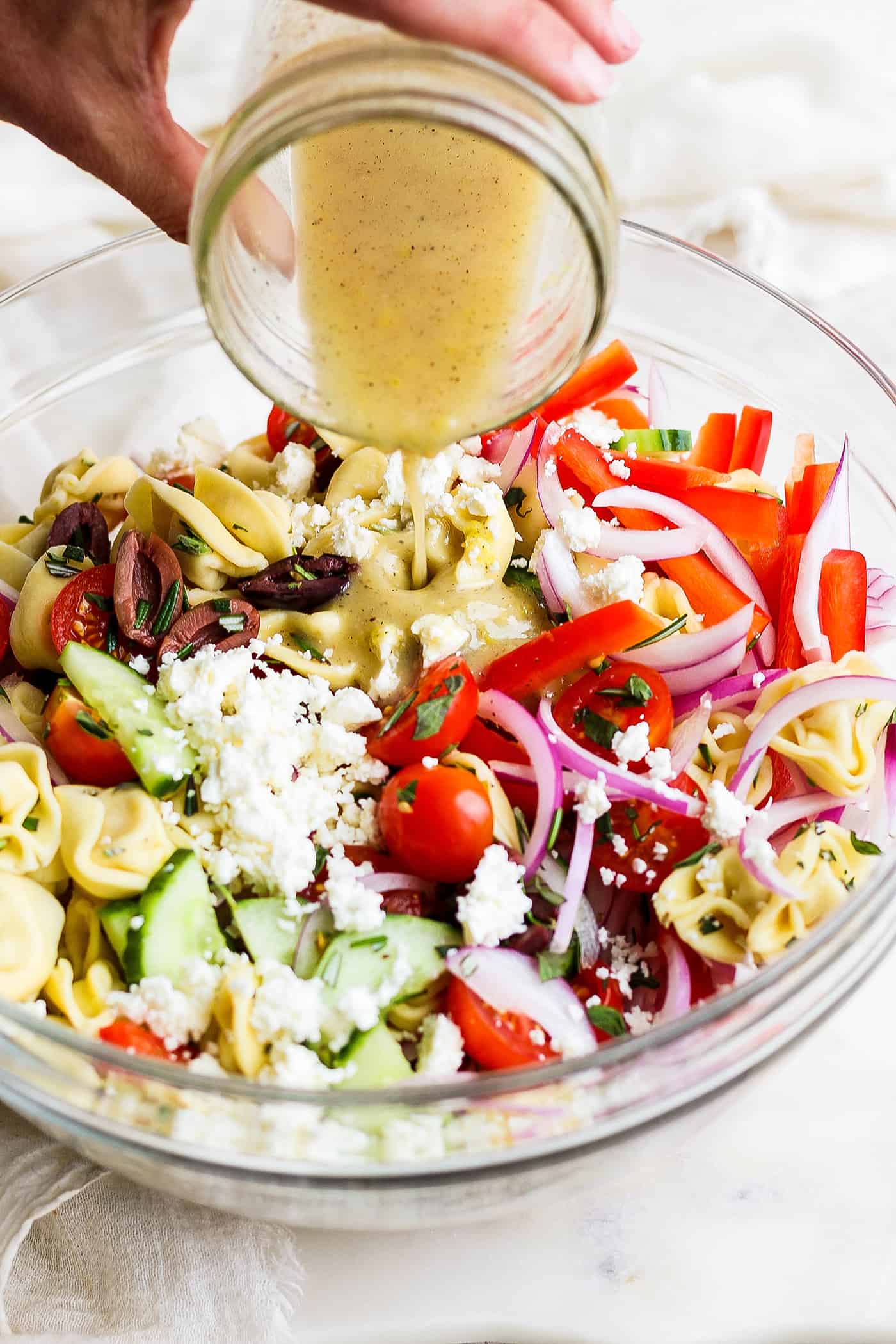 A hand pours dressing onto a bowl of Greek tortellini salad.