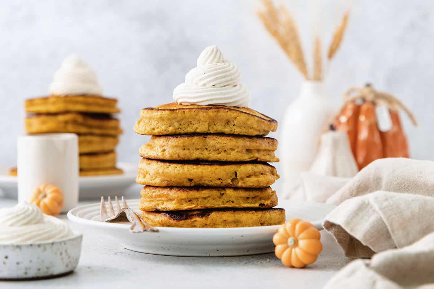 A stack of pumpkin pancakes topped with maple cinnamon whipped cream on a white plate.