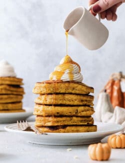 Maple syrup is poured over a stack of pumpkin pancakes with mini pancakes scattered around the plate.