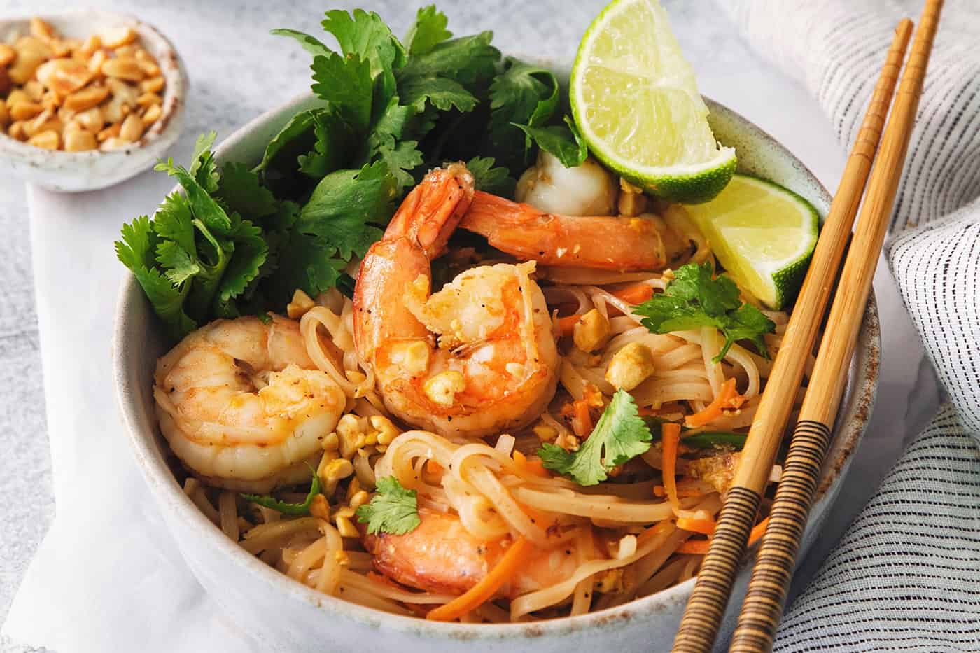 A bowl of shrimp pad thai in a bowl with lime wedges and chopsticks along the edge of the bowl.