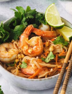 A bowl of shrimp pad thai in a bowl with lime wedges and chopsticks along the edge of the bowl.