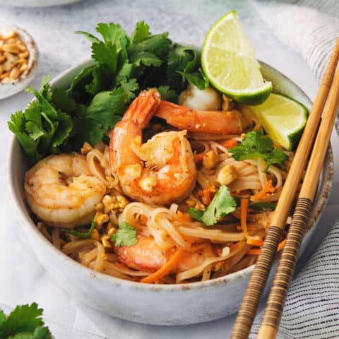 A bowl of shrimp pad thai in a bowl with lime wedges and chopsticks along the edge of the bowl.