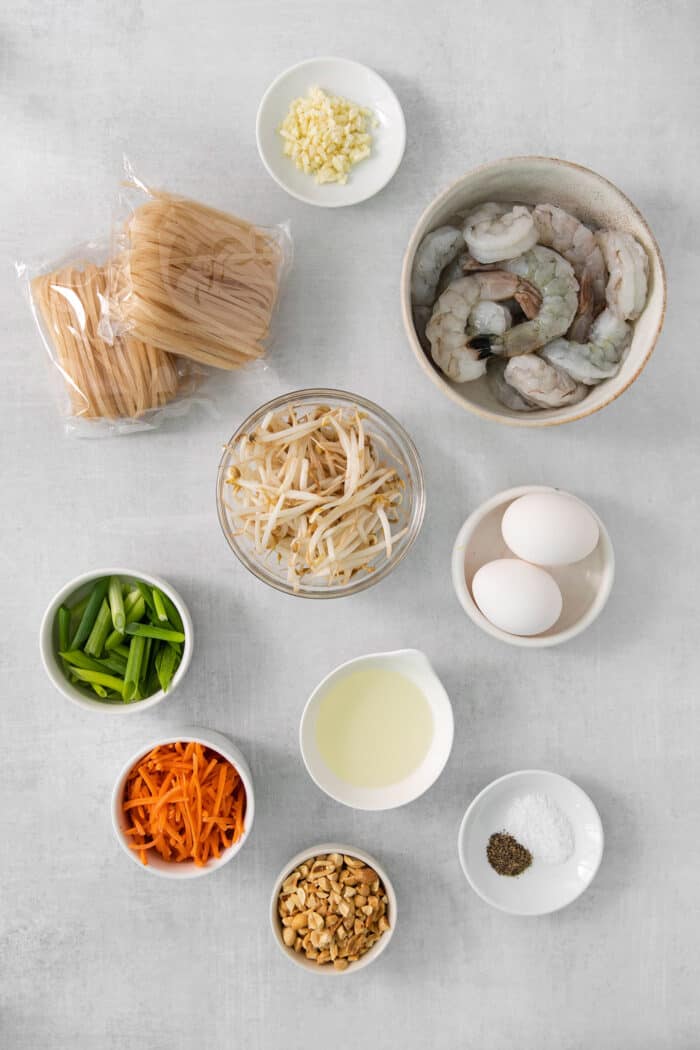 The ingredients for shrimp pad Thai are shown in bowls on a white background: shrimp, green onions, bean sprouts, carrots, eggs, oil, salt, and noodles.