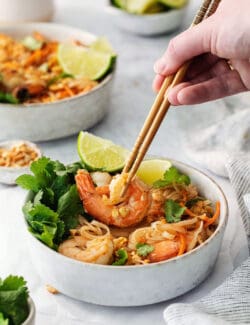 A hand holding chopsticks lifts out a piece of shrimp from a white bowl of shrimp pad Thai with more bowls in the background.