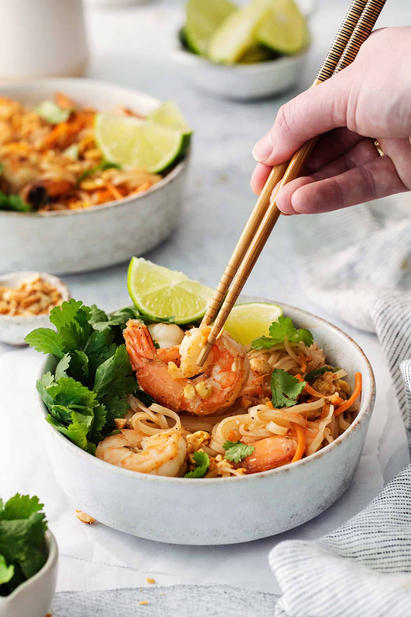 A hand holding chopsticks lifts out a piece of shrimp from a white bowl of shrimp pad Thai with more bowls in the background.