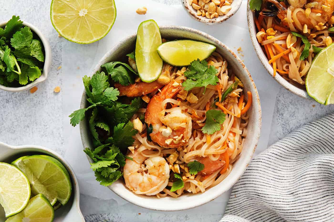 An overhead shot of a bowl of shrimp pad Thai on a white background surrounded by bowls of garnish.