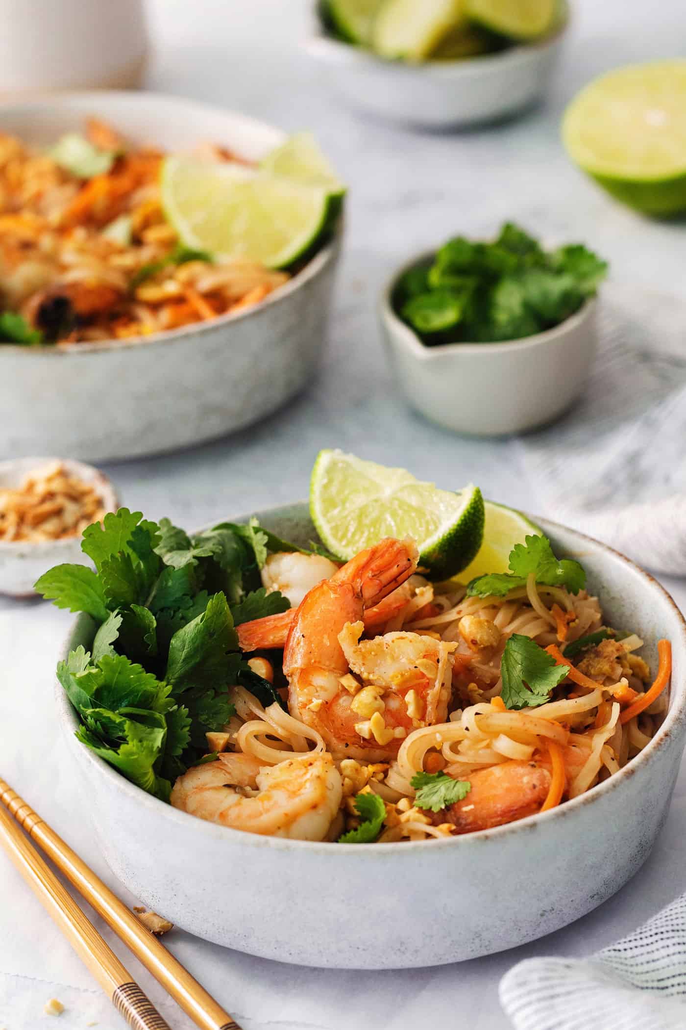 A table with white bowls of shrimp pad Thai, chopsticks, and lime.