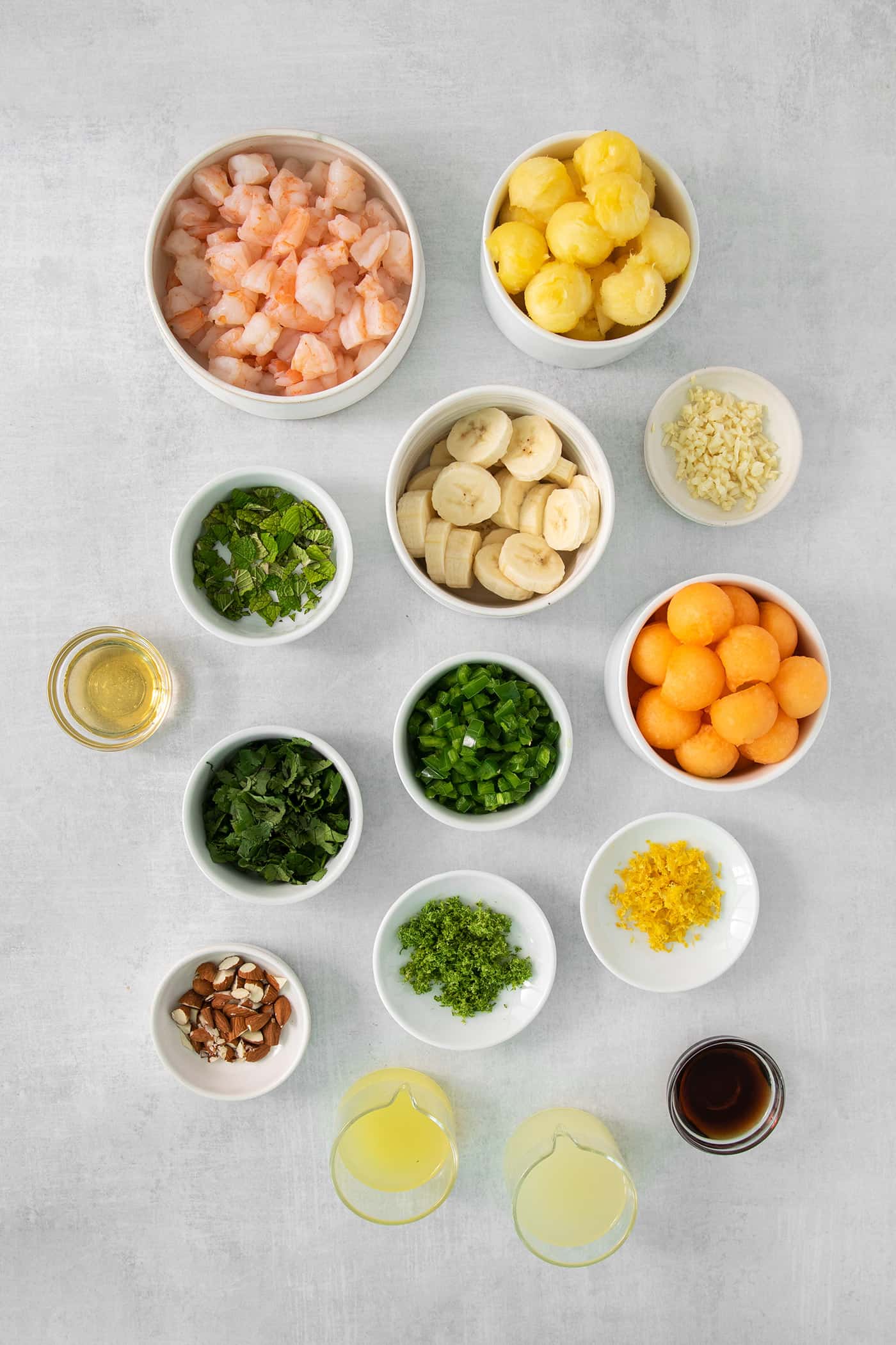 The ingredients needed to make tropical shrimp salad are shown in bowls, including melon balls, pineapple, banana slices, green onions, mint and cilantro, shrimp, and almonds.