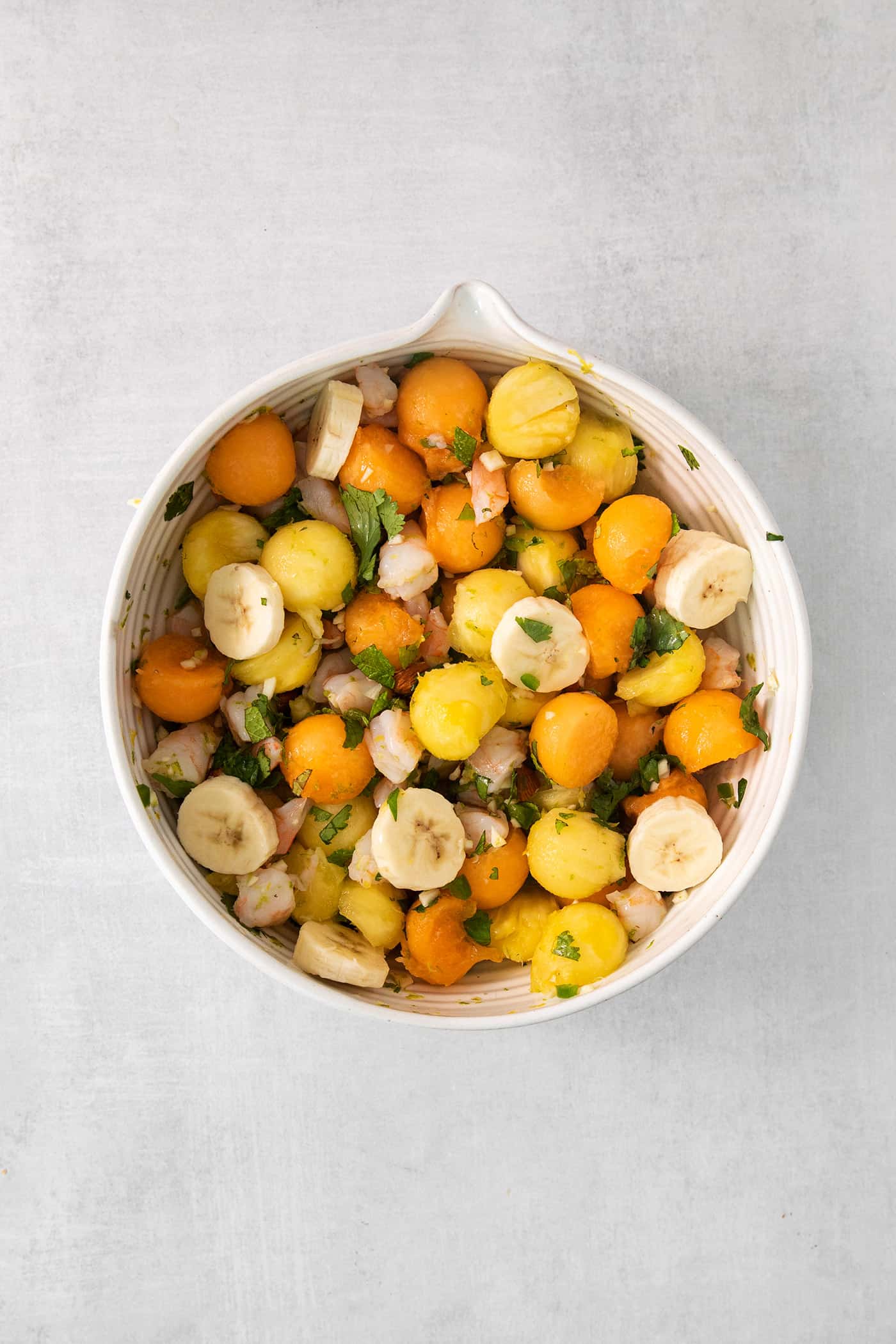 Melon, banana, and pineapple is added to a white bowl of tropical shrimp salad, shown on a white background.