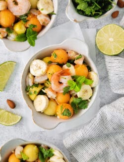 An overhead shot of colorful bowls of tropical shrimp salad with fruit, shrimp, and mint leaves and limes on the side.