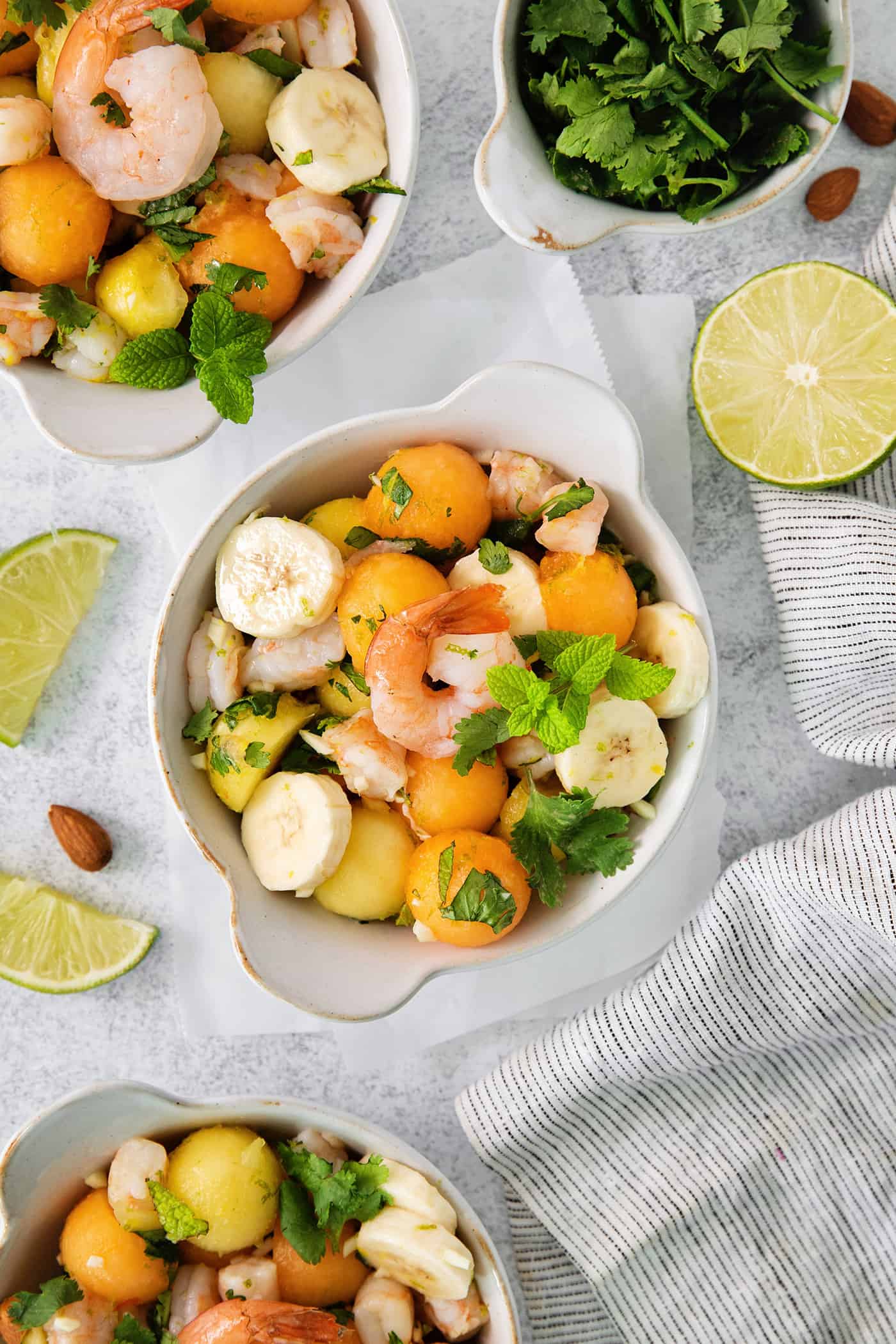 An overhead shot of colorful bowls of tropical shrimp salad with fruit, shrimp, and mint leaves and limes on the side.