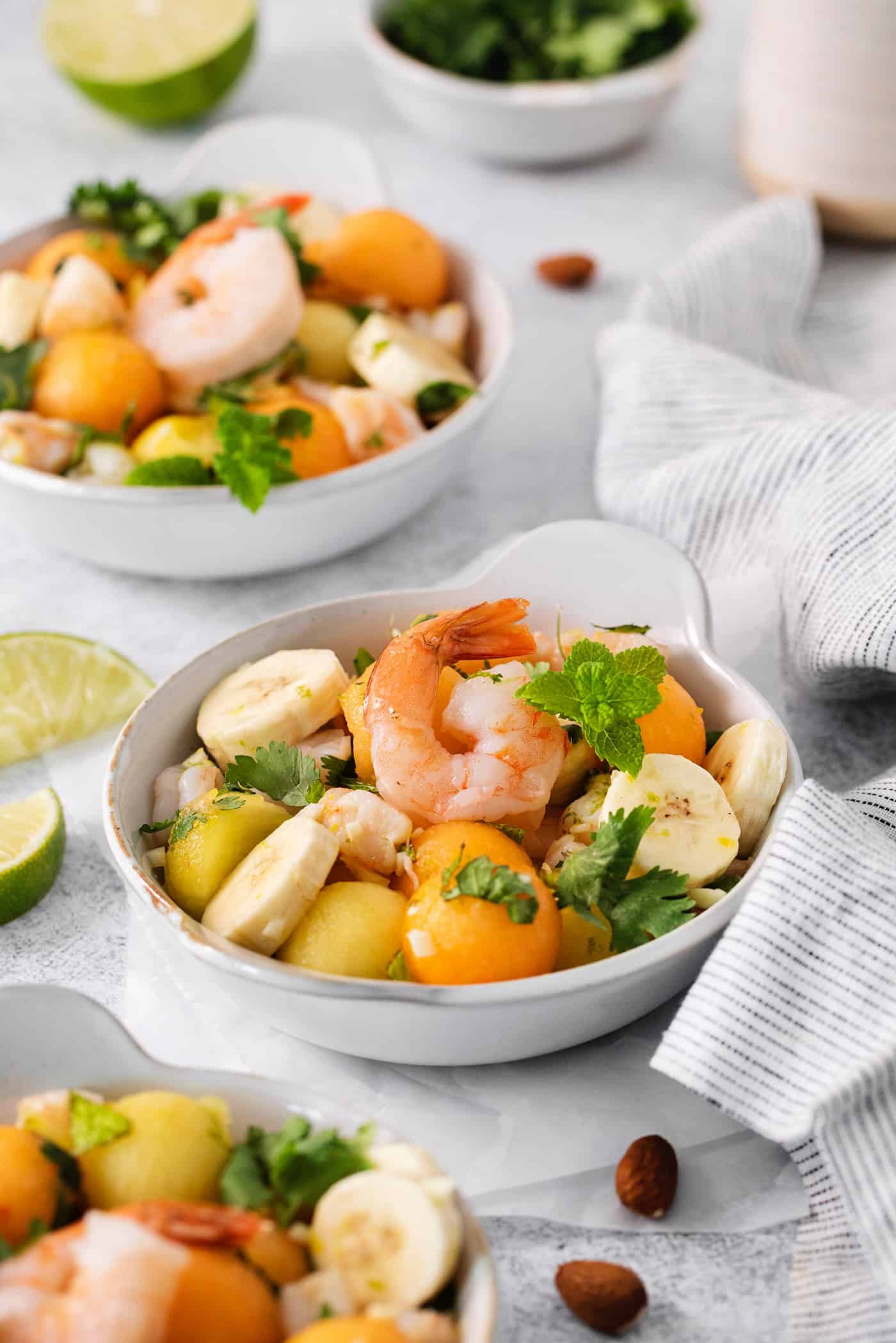 Colorful bowls of tropical shrimp salad with fruit, shrimp, and mint leaves and limes on the side are shown on a table with napkins.