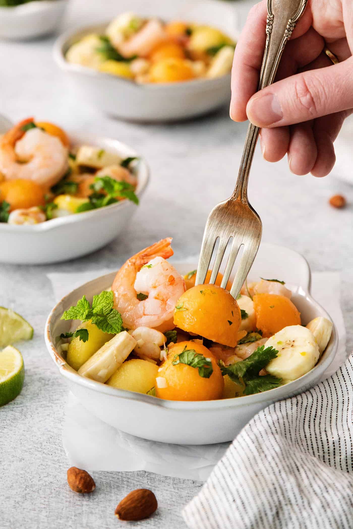 A hand holding a fork picks up a piece of melon out of a bowl of tropical shrimp salad with another bowl of salad shown in the background.